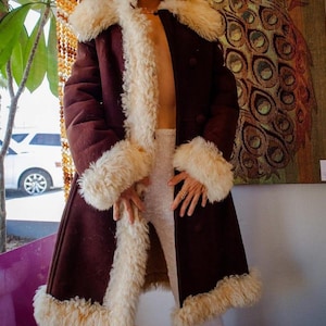 Op de afbeelding: Een lange, donker bordeauxrode jas met crèmekleurige imitatiebontafwerking op de kraag, manchetten en zoom. De jas heeft een dubbelborstig ontwerp en een vintage esthetiek. Het model draagt een crèmekleurige outfit.