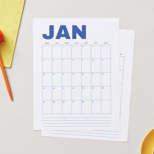 Op de afbeelding: Een stapel witte papieren kalenders met de maand januari in blauw gedrukt. Een gele notitieblok met een pomponpen en een gele beker zijn ook zichtbaar.