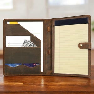 Puede incluir: Portafolio de cuero marrón abierto con múltiples bolsillos, incluyendo ranuras para tarjetas y efectivo. El portafolio contiene un bloc de notas con papel rayado amarillo y un portalápices. El cuero tiene un acabado envejecido y el portafolio está abierto sobre una superficie de madera.