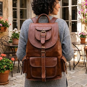 May include: A brown leather backpack featuring a top flap with a buckle closure, a vertical strap with a buckle, and two side pockets. The bag has a top handle and adjustable shoulder straps, constructed with visible stitching and brass-toned hardware.
