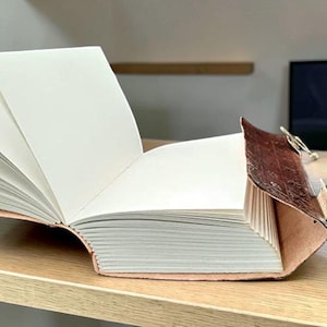 May include: A leather-bound journal with blank white pages, open to display the interior. The cover is brown with a textured finish, fastened with a metal clasp. The journal sits on a light-coloured wooden surface, possibly a desk.