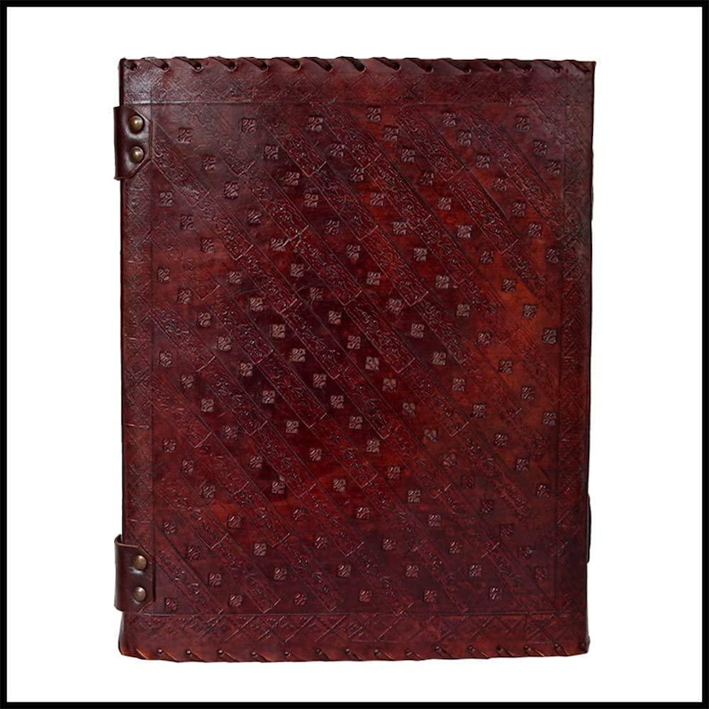 May include: A dark brown leather journal with an embossed pattern. The cover has a diagonal design of small squares and lines. The edges are stitched, and the journal features metal corner accents and a leather strap closure. The journal measures approximately 25 cm x 20 cm.