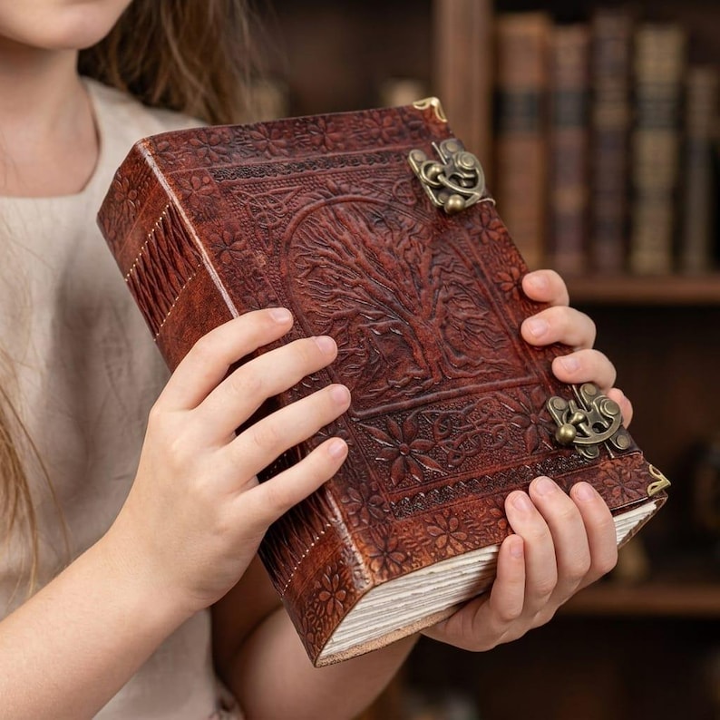 May include: A brown leather journal with an embossed tree design on the cover. The journal has a clasp closure and decorative corner pieces. The pages are visible, and the journal is held by a person.