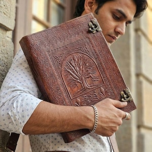 May include: A brown leather journal with an embossed tree design on the cover. The journal has decorative metal clasps and is held by a person. The journal is a rich brown colour and appears to be handmade.
