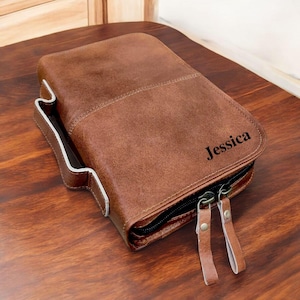 May include: A brown leather case with the name "Jessica" embossed on the front. The case has a zip closure with two leather pull tabs and a white-lined pocket on the side. The case is on a wooden surface.
