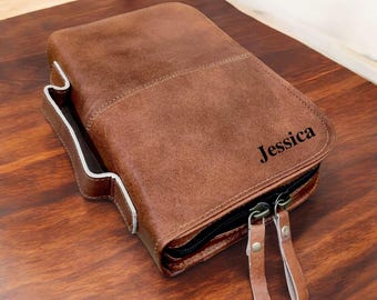 Puede incluir: Un estuche de cuero marrón con el nombre "Jessica" grabado en la parte delantera. El estuche tiene un cierre de cremallera con dos tiradores de cuero y un bolsillo forrado en blanco en el lateral. El estuche está sobre una superficie de madera.