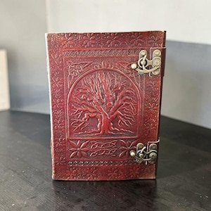 May include: A burgundy leather journal with an embossed tree of life design on the cover. The journal has a decorative floral border and a metal clasp. The cover is a rich, reddish-brown colour.