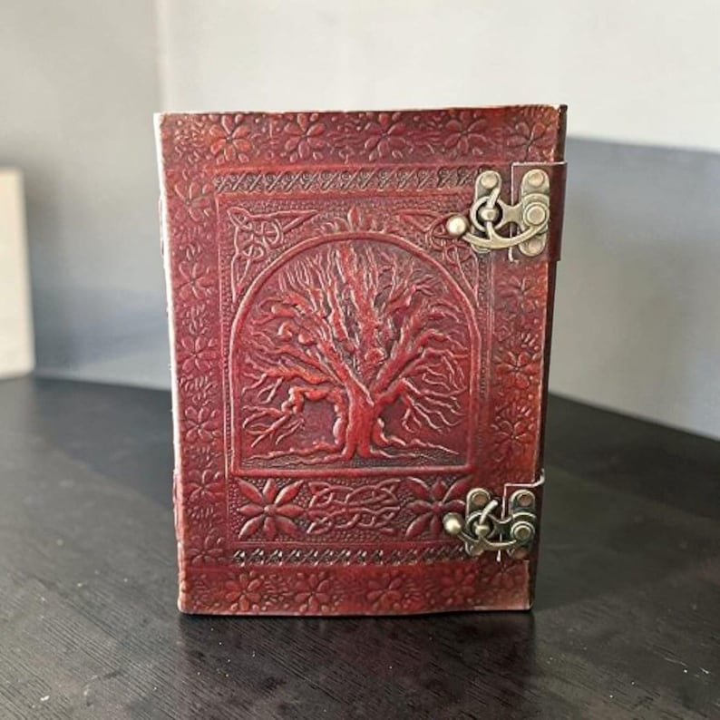 May include: A burgundy leather journal with an embossed tree of life design on the cover. The journal has a decorative floral border and a metal clasp. The cover is a rich, reddish-brown colour.