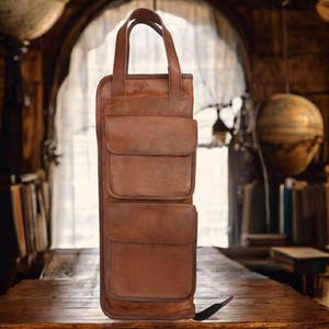 May include: A brown leather wine bottle carrier with two front pockets and a top handle. The carrier is designed to hold a single bottle and is made of a rich, textured leather. The carrier is approximately 14 inches tall.