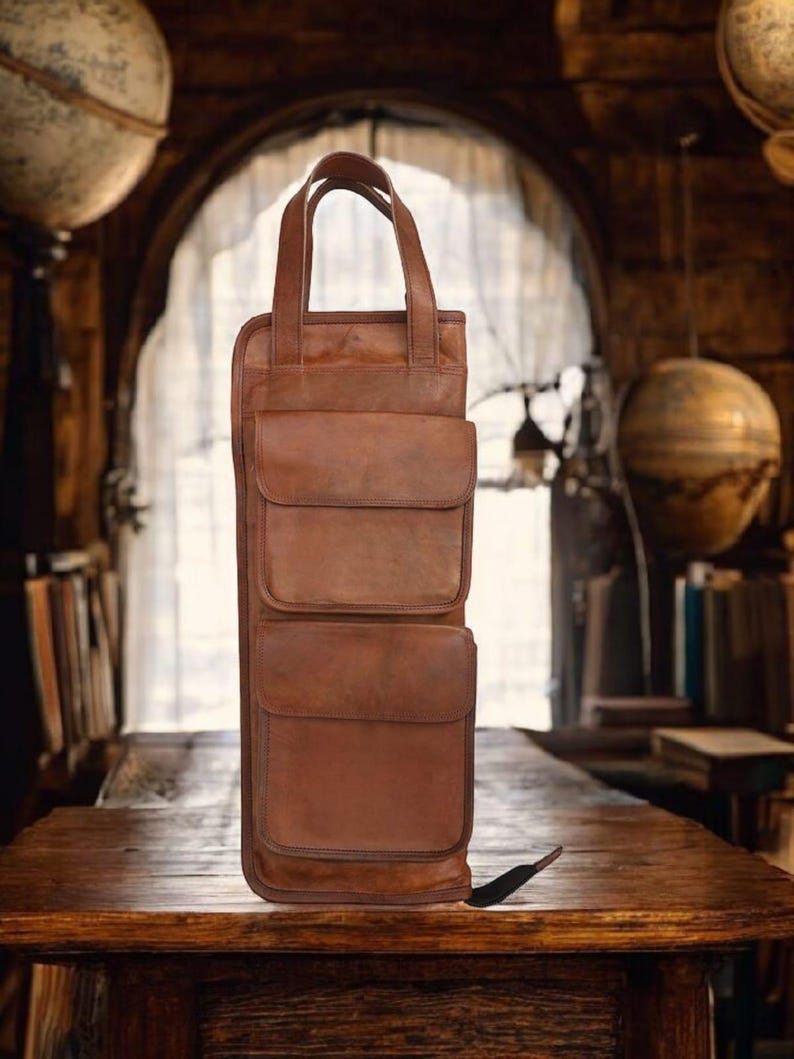 May include: A brown leather wine bottle carrier with two front pockets and a top handle. The carrier is designed to hold a single bottle and is made of a rich, textured leather. The carrier is approximately 14 inches tall.