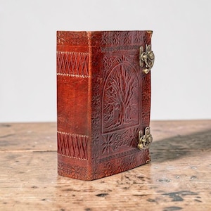 May include: A brown leather journal with an embossed tree of life design on the cover. It features a clasp closure and decorative stitching on the spine. The journal is on a wooden surface.