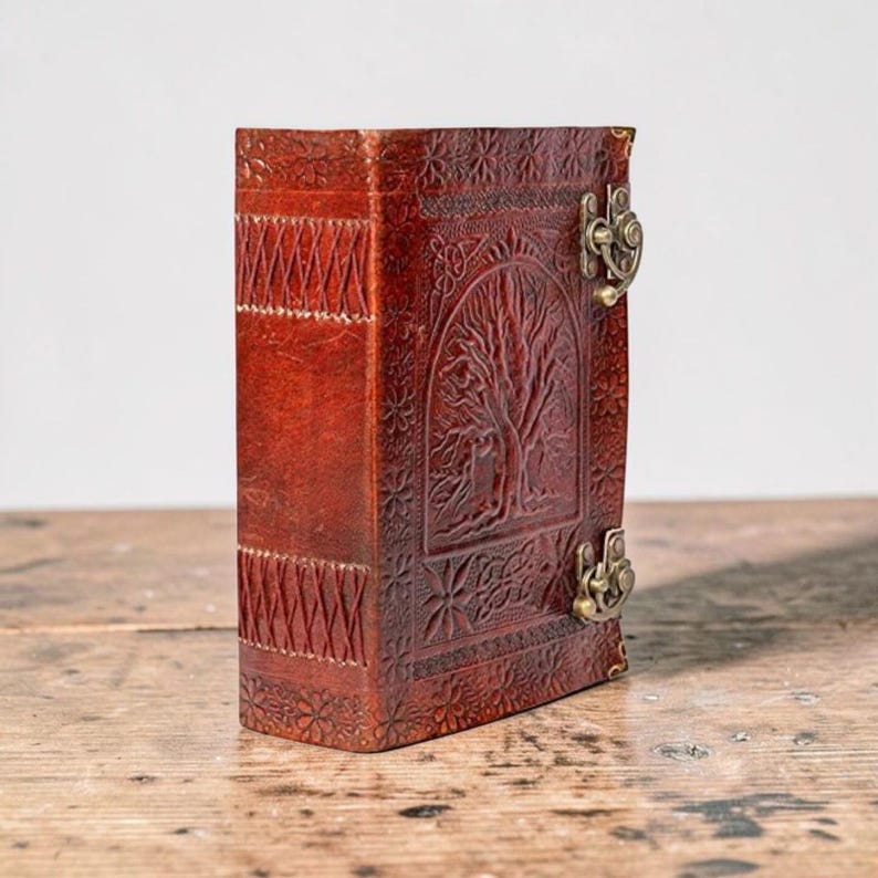 May include: A brown leather journal with an embossed tree of life design on the cover. It features a clasp closure and decorative stitching on the spine. The journal is on a wooden surface.