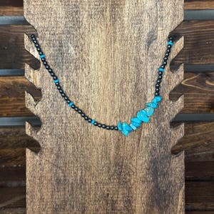 May include: A necklace with black beads and turquoise stone accents displayed on a wooden jewelry stand. The necklace features a strand of small black beads with interspersed turquoise beads and larger turquoise stone pieces.