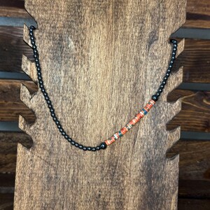 May include: A black beaded necklace with a section of orange, teal, and white beads. The necklace is displayed on a wooden jewelry stand with a dark brown finish. The stand has a decorative, notched design.