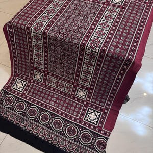 Puede incluir: Un textil rectangular con un fondo granate profundo y complejos patrones geométricos y florales en blanco y negro. El diseño incluye motivos cuadrados repetitivos, bordes con elementos circulares y una banda negra sólida en el borde inferior.