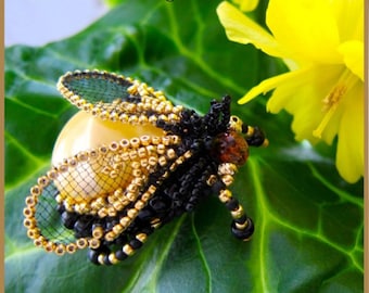 May include: A beaded bug, crafted with black and gold beads, rests on a green leaf. The bug's wings are a delicate mesh, and the body is detailed with intricate beadwork. The image includes text that reads "PDF Tutorial The Beaded Bug" and "Made in USA".