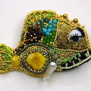 May include: A whimsical beaded fish brooch with a large amber stone, turquoise and green beads, and a pearl accent. The fish has a large, white eye with a black pupil and eyelashes.