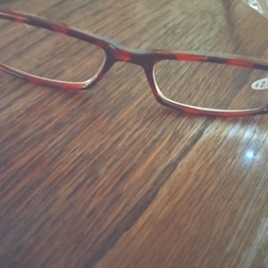 May include: Rectangular reading glasses with a tortoiseshell pattern frame. The lenses have a +2.50 magnification. The glasses are resting on a wooden surface.