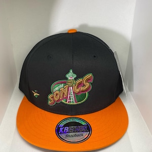 May include: Black and orange Seattle Sonics snapback hat. The hat features the team logo on the front, with the words "Seattle Sonics" in green and gold. The brim is orange, and the top of the hat is black.