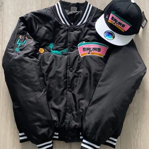 May include: Black satin bomber jacket and baseball cap set. The jacket displays the San Antonio Spurs logo in pink, teal, and white. The cap has a white peak and the Spurs logo. The jacket has white and black striped cuffs and collar.