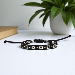 Puede incluir: Pulsera de macramé negra con cuentas plateadas. La pulsera presenta un diseño tejido con cuadrados negros y plateados alternos. Cordones negros ajustables se extienden desde cada extremo de la pulsera.