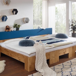 Op de afbeelding: Een tweepersoonsbed met een licht houten frame, blauw hoofdbord en witte beddengoed. Het bed heeft een gestreepte deken en zachte kussens. Een houten nachtkastje met een lamp en decoratie staat naast het bed. Een zacht tapijt ligt op de vloer.