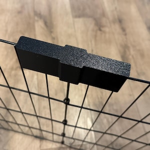 May include: A black metal wire panel with a black plastic connector. The connector is rectangular with a textured surface and is attached to the wire panel. The background is a light brown wooden surface.