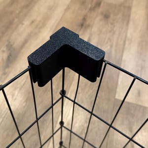 May include: Black corner bracket attached to a black wire mesh basket. The bracket is textured and L-shaped, designed to fit the corner of the basket. The basket has a grid pattern, and the background is a light brown wooden surface.