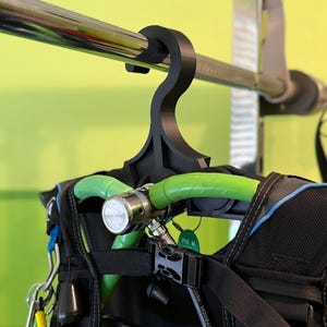 May include: A black scuba diving BCD (Buoyancy Control Device) hanging from a black hanger on a metal bar. The BCD has green hoses and silver metal components. The background is a light green wall.