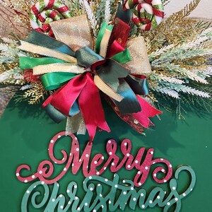 May include: A green Christmas ornament with the words "Merry Christmas" in red and white polka dots. The ornament is decorated with a bow, candy canes, and greenery.