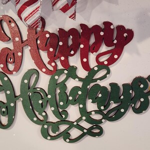 May include: A festive holiday decoration featuring the words "Happy Holidays" in red and green, with white polka dots. The words are on a white background, accented with ribbons in red and white candy cane stripes, green, and silver.