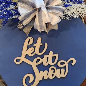 May include: A round, navy blue wooden sign with the phrase "Let it Snow" in a cursive font. The sign is decorated with a white bow, silver and gold ribbons, and blue and white floral accents.