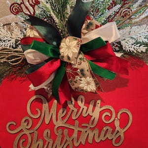 May include: A red wooden Christmas ornament with the words "Merry Christmas" in gold script. The ornament is decorated with greenery, white accents, and bows in red, green, and cream.