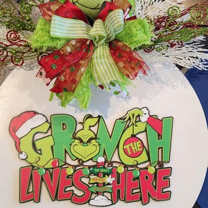 May include: A round, white Grinch-themed sign with the text "Grinch Lives Here" in green and red. The sign is decorated with a Grinch plush head wearing a Santa hat, bows, and decorative greenery.