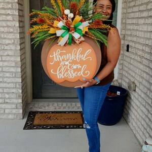 May include: A round wooden pumpkin-shaped sign with the words "Thankful & Blessed" in cursive script. The sign is decorated with fall foliage, ribbons, and small pumpkins. A festive autumn decoration.