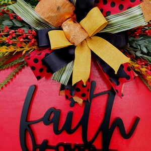 May include: A round, red wooden sign with the words "faith over fear" in black script. The sign is decorated with a variety of colorful ribbons, bows, and faux greenery, including yellow, black, and red polka dot patterns.