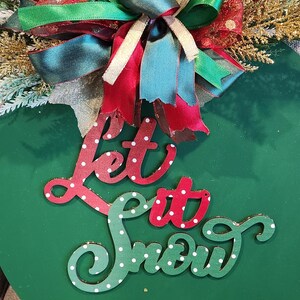 May include: A round, green Christmas ornament with the words "Let it Snow" in red and green polka-dotted lettering. The ornament is decorated with a large bow of red, green, and gold ribbons and sprigs of greenery.