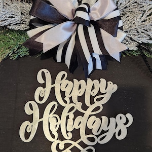 May include: A round, black holiday decoration with a silver "Happy Holidays" script. It features a bow with black, white, and silver ribbons, and is adorned with silver and green decorative sprigs. A festive seasonal accent.
