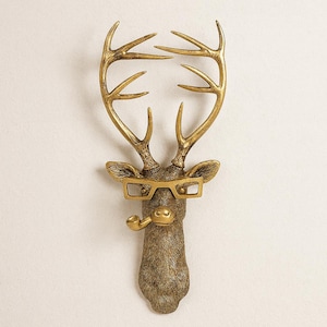 May include: A decorative deer head sculpture with gold-colored antlers, glasses, and a pipe. The sculpture is brown and gold, with the text "The Animal Objet" in a script font.