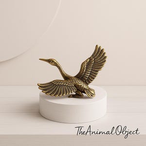 May include: A bronze-coloured metal sculpture of a bird with outstretched wings, perched on a white cylindrical pedestal. The bird's feathers are intricately detailed, and its beak is pointed. The sculpture is a decorative object.