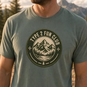 May include: A sage green short-sleeve t-shirt featuring a circular graphic on the chest. The design includes a mountain range, a forest, and a lake, surrounded by the text "TYPE 2 FUN CLUB" with small star and pine tree icons in white.