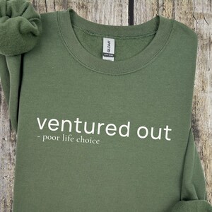 May include: An olive green sweatshirt with the words "ventured out - poor life choice" printed in white. The sweatshirt is made of a soft material and has a crew neck. The Gildan brand tag is visible.