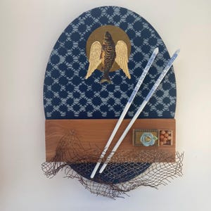 May include: A wall-mounted art piece featuring an oval dark blue panel with a white geometric pattern, a central gold circle with a winged fish, a horizontal wooden plank, two white chopsticks, a small floral ornament, and a metal mesh overlay.