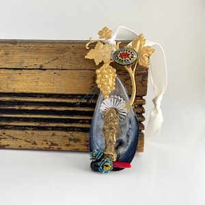 May include: A gold and blue pendant featuring a religious figure, a grapevine, and a red coral accent. The pendant is attached to a white ribbon.