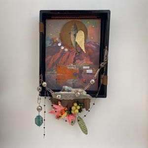 May include: A black shadow box frame containing a collage of a fish with a gold wing, a ship, and mountains. A silver chain with beads, charms, and a green pendant hangs from the bottom. Artificial leaves and yellow berries are attached below.