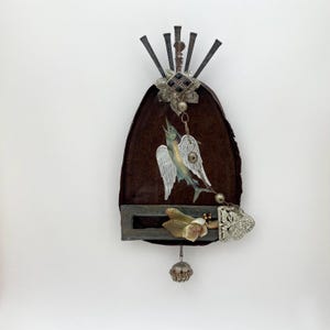 May include: A mixed media wall hanging with a dark brown, bell-shaped base. It features a painted fish with angel wings, metal embellishments, and a decorative floral element. The piece has a vintage, handcrafted aesthetic.