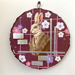 May include: A round wooden plaque with a white rabbit wearing a patterned jacket, surrounded by pink and white flowers and silver geometric shapes.