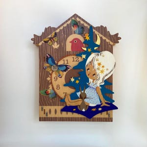 May include: A whimsical wooden cuckoo clock featuring a girl, a rabbit, and a blue Christmas tree. The clock face is tan with black numbers. Butterflies and a red bird are also featured in the design. The clock is mounted on a white wall.