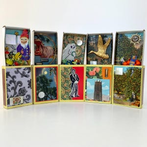 May include: A collection of decorated matchboxes, each with unique artwork and embellishments. The boxes feature images of a gnome, a hippo, a bride, a golden duck, and a landscape. The bottom row displays patterned designs and illustrations.