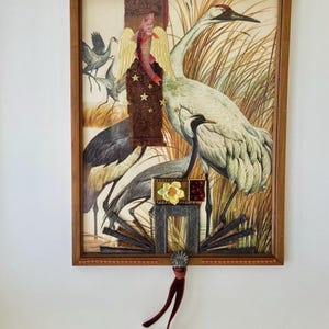 May include: A framed mixed-media collage featuring illustrations of large white and grey cranes in tall grass. The artwork includes a central winged fish, a small yellow flower, dark red beads, a metal arch, and a dangling red velvet ribbon.